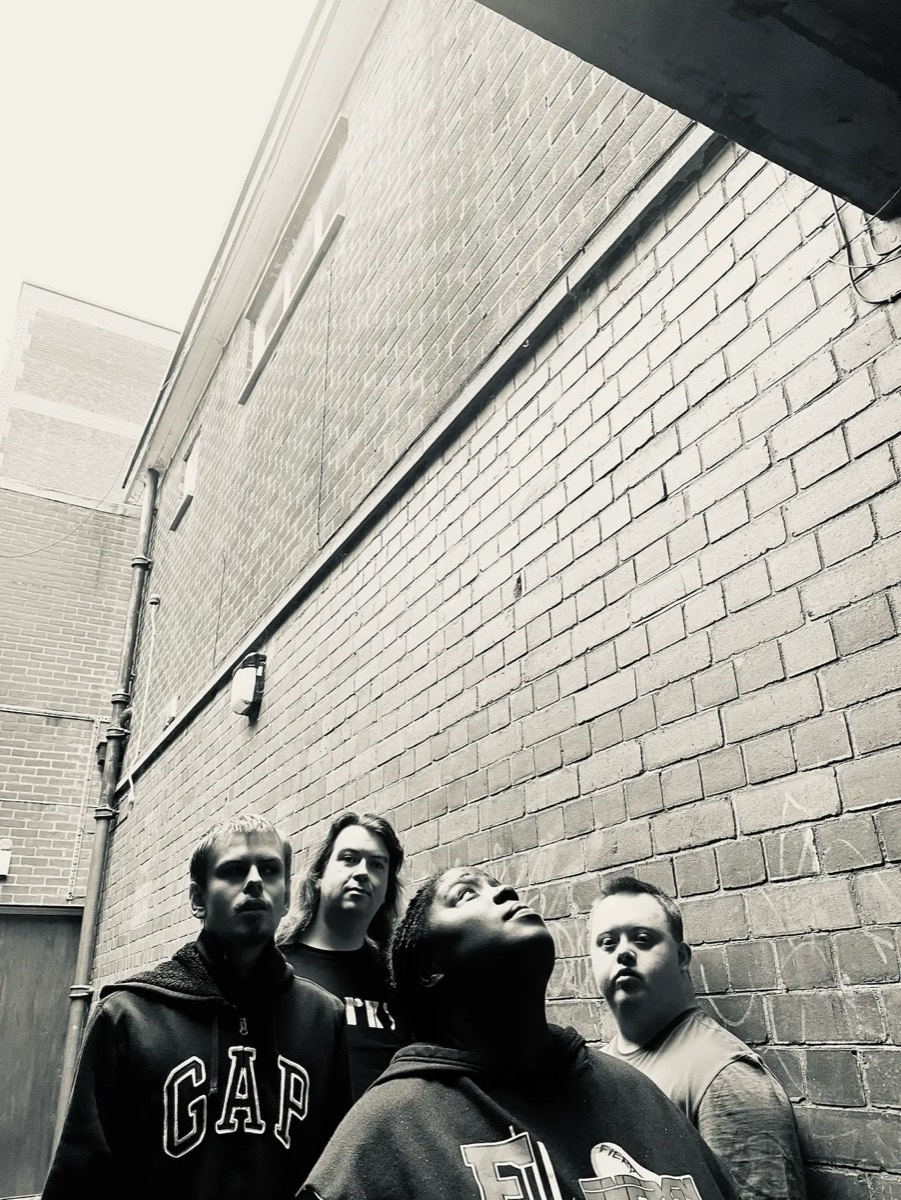 The four members of Little Trees in black and white, standing against a tall brick wall in a narrow alley. Shimara, at the front, is looking up at the sky; the other three look towards the camera.